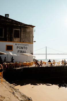 People enjoy dining at the riverside Ponto Final in Lisbon with bridge view and sunny weather.