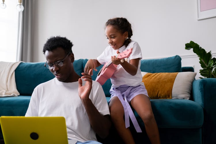 A Young Girl Sitting On The Couch While Disturbing Her Father Working On Laptop