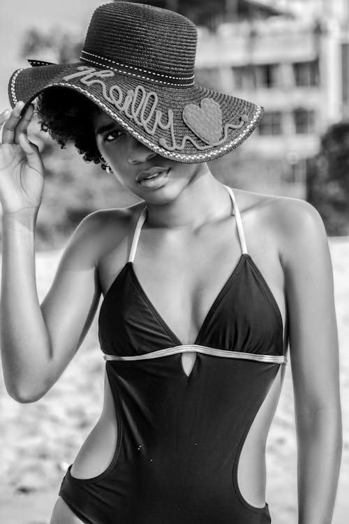 Black and white of slim African American female in swimsuit and trendy hat looking at camera on beach in summer
