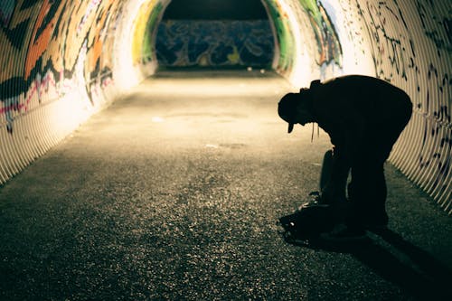 Side view of anonymous male silhouette with backpack leaning forward in tunnel with graffiti on walls in evening town