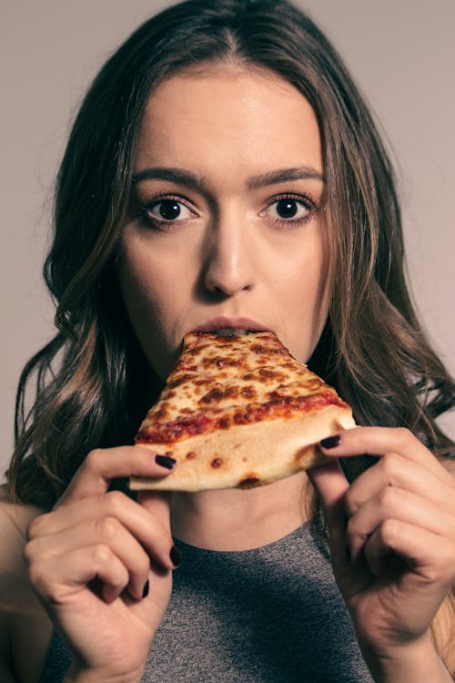 Astonished young female with long dark biting slice of tasty pizza and looking at camera in studio
