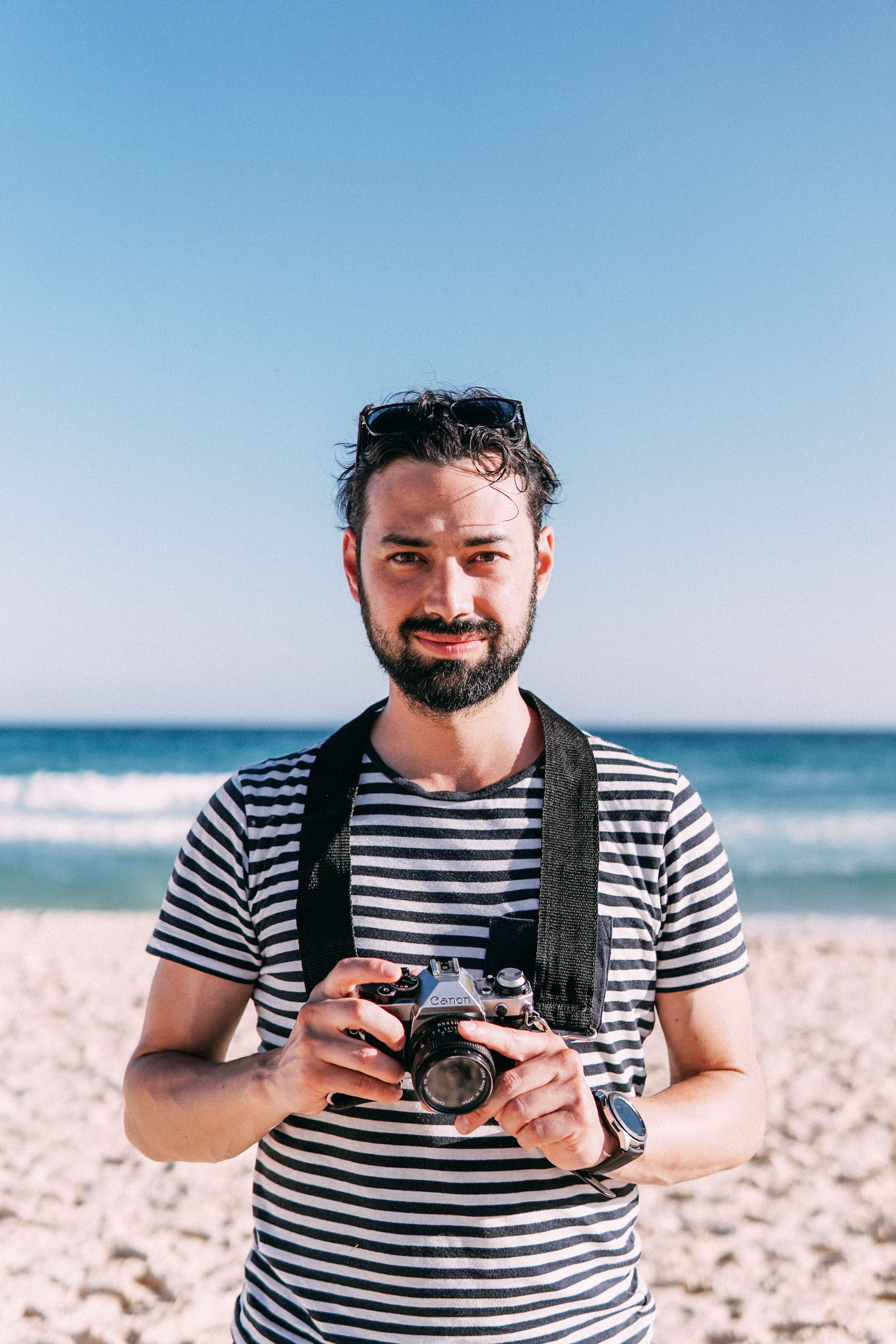 Portrait of a bearded man holding a camera on a sunny beach day.