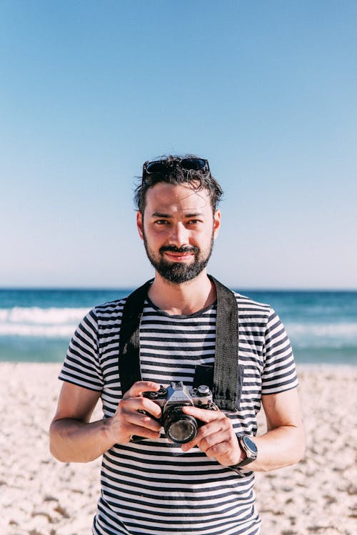 A Man in Striped Shirt Holding DSLR Camera while on the Beach