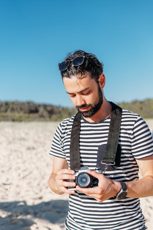 A Man in Striped Shirt Holding DSLR Camera while on the Beach