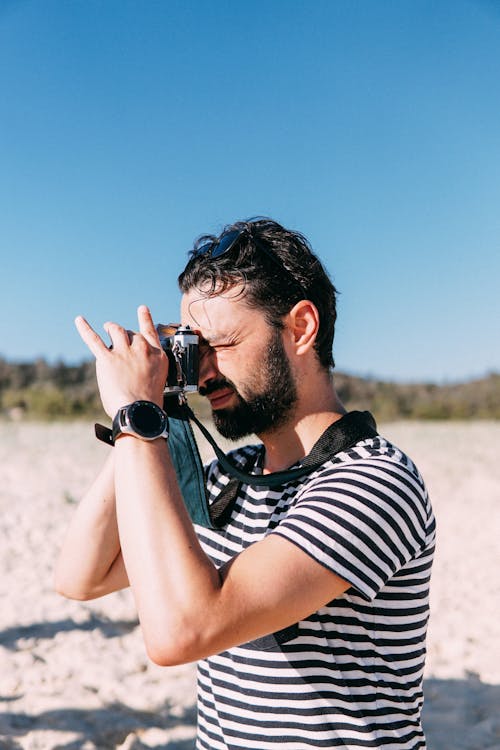 A Man in Striped Shirt Using DSLR Camera while on the Beach