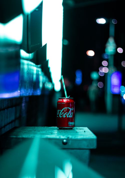 Free Coca Cola can with straw on a bench, bokeh neon lights in London street at night. Stock Photo
