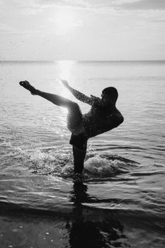 Silhouette of a male kickboxer performing a high kick in the water during sunset.
