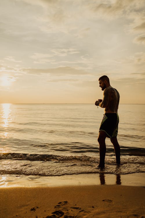 Man in Black Shorts Standing on the Seashore During Sunset
