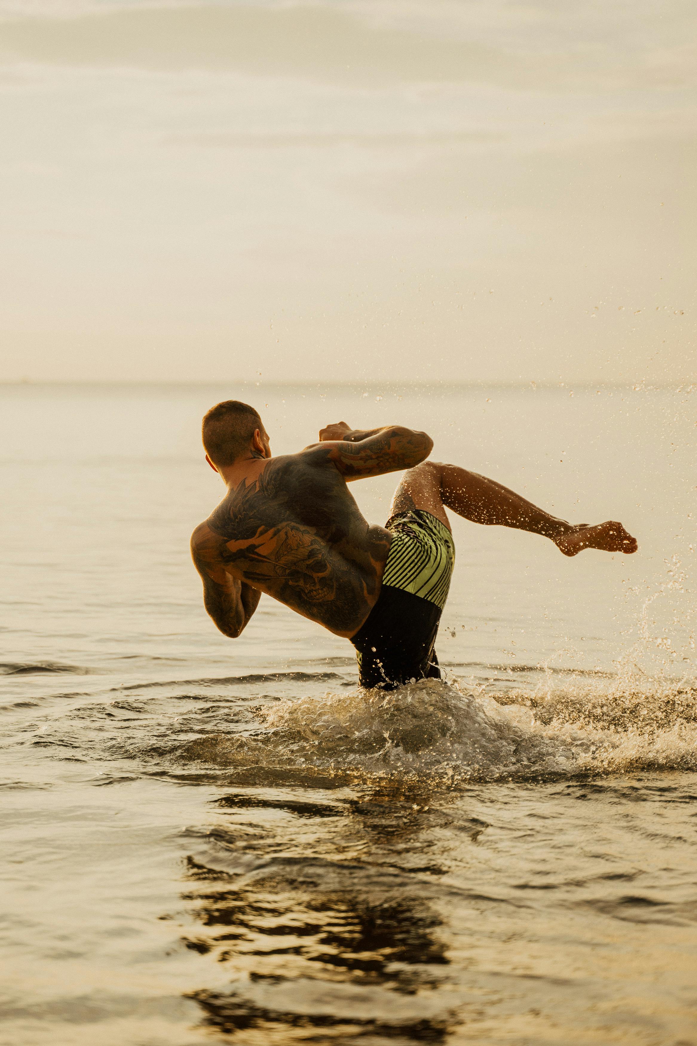 A man performs powerful martial arts kicks in the water at sunset, showcasing skill and strength.