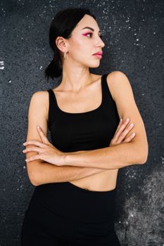 一位自信的女士身穿运动服，双臂交叉，倚靠在有纹理的黑色墙壁上
