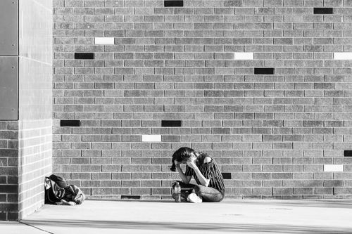Free Black and white photo of a student sitting alone by a brick wall, deep in thought. Stock Photo
