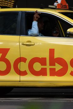 A taxi driver in a yellow cab talking on a phone while driving through a bustling urban area.