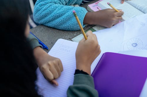 Crop faceless ethnic children sitting at table with pencils and doing homework together