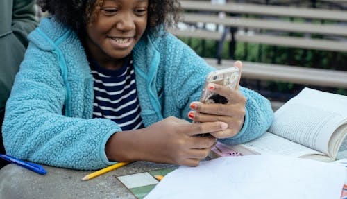 Crop African American child smiling and browsing mobile phone while having break from homework
