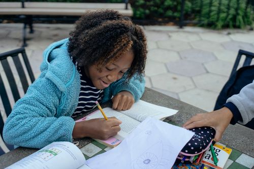 Free Happy African American child in casual clothes sitting at table with pencil and writing homework while spending time with classmate Stock Photo