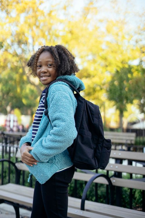 Free Side view of cheerful black little girl with backpack standing in park near wooden bench and looking over shoulder Stock Photo