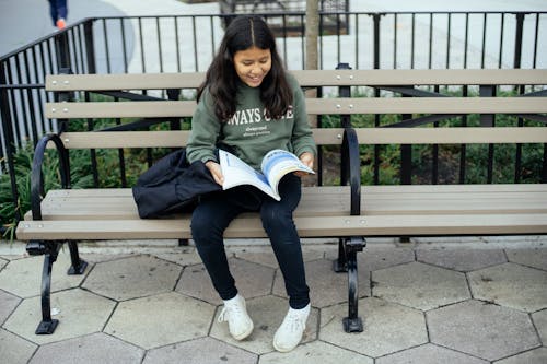 Cheerful Hispanic girl with textbook on bench