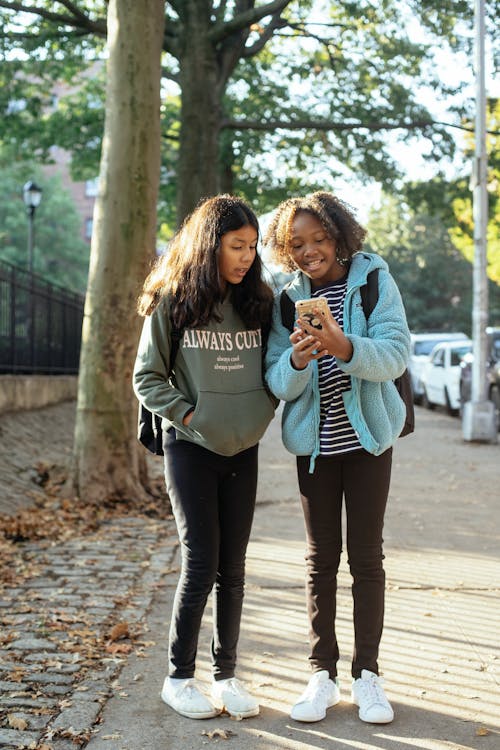 Cheerful schoolgirls with smartphone on sidewalk