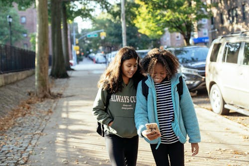 Cheerful diverse schoolgirls with backpacks walking on city street while browsing mobile phone together