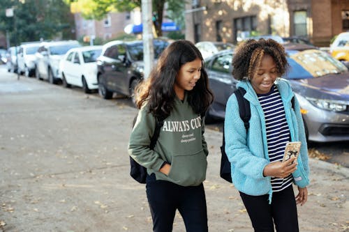 Cheerful multiracial little girls with backpacks strolling together and browsing mobile phone on sidewalk