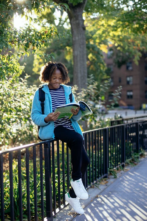 Free Full length of little African American girl in casual clothes sitting on metal fence with backpack and studying school task Stock Photo