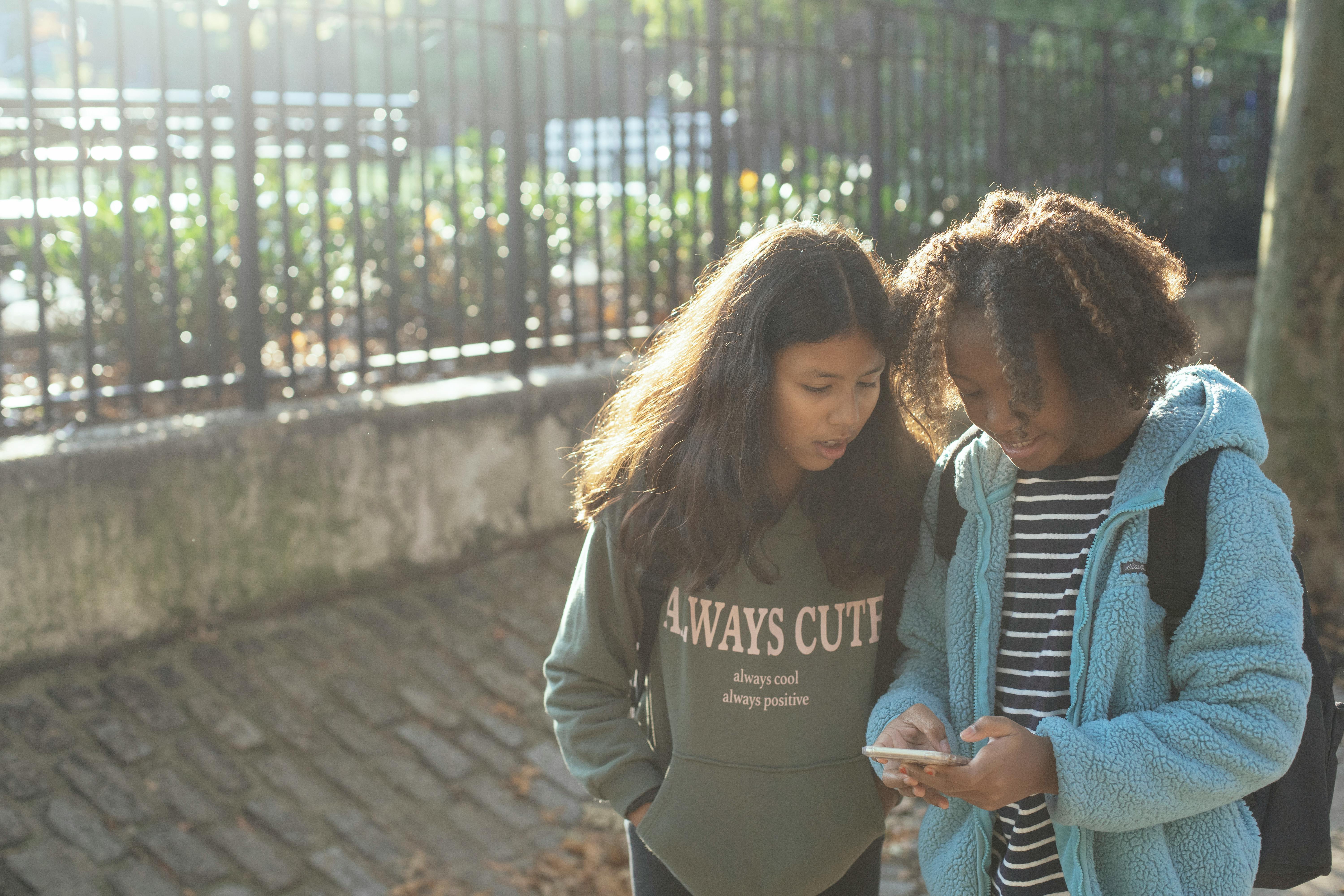 excited multiethnic schoolgirls browsing smartphone on sidewalk