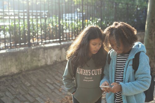 Little diverse girls in casual clothes with backpacks standing near park and browsing mobile phone in sunny day