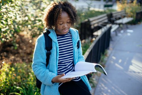 Free African American child in casual clothes with backpack sitting on metal fence and reading book while spending time in park Stock Photo
