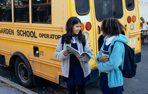Free Happy multiracial classmates with backpacks standing near yellow school bus and communicating after school while looking at each other Stock Photo