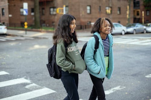 Content multiracial girlfriends with school bags and hands in pockets interacting on urban crosswalk while looking away