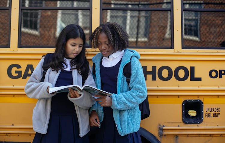 Multiracial Schoolgirls With Workbook Near Bus In City