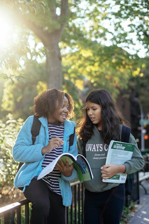 Cheerful diverse schoolgirls with workbooks talking on street