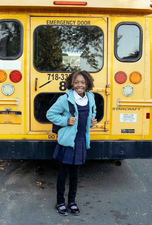 Content African American schoolkid in uniform looking at camera near bright bus on urban asphalt roadway