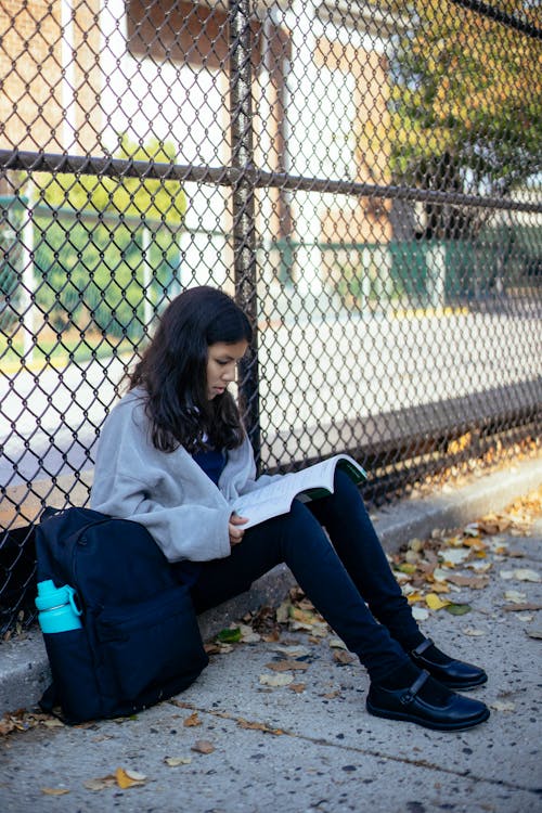 Ethnic schoolgirl with workbook studying near grid fence and backpack