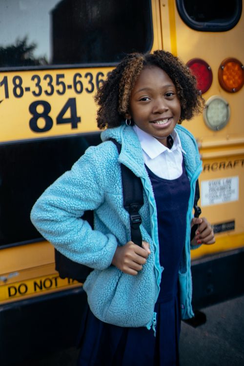 Free Cheerful African American kid with school bag looking at camera near bright bus in city Stock Photo