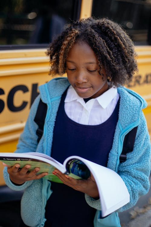 Positive black girl reading textbook near school bus