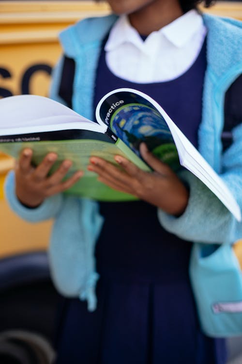 Crop unrecognizable schoolgirl reading textbook while standing near yellow school bus on sunny autumn day