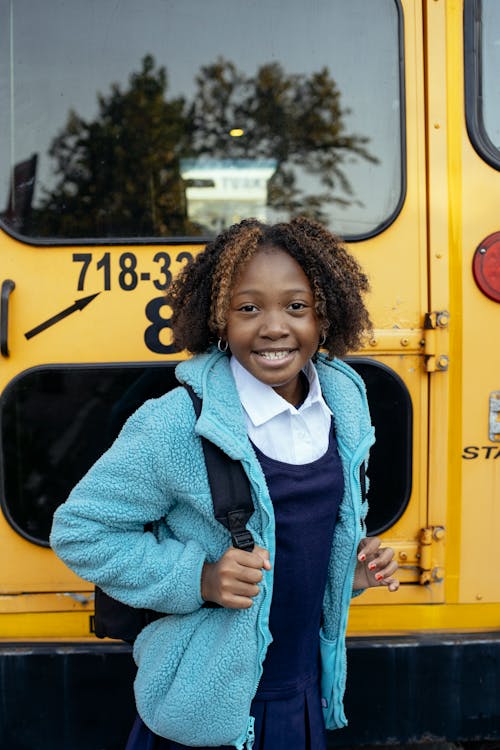 Cheerful African American girl with curly hair standing with backpack near yellow school bus and looking at camera with smile