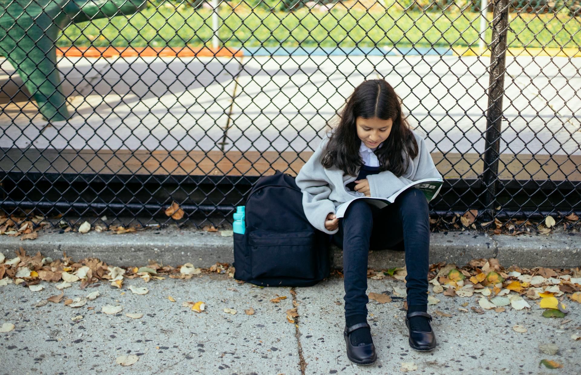 Full body concentrated schoolgirl in warm sweater reading topic in textbook while sitting on ground near net fence in autumn park