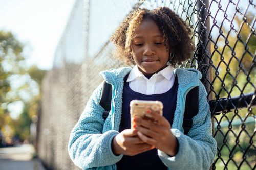 Free Positive African American schoolgirl in blue  jacket browsing mobile phone while standing near wire net fence on sunny autumn day Stock Photo