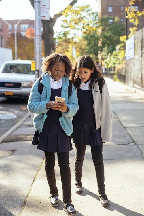Full body positive multiethnic girls classmates walking on sunny autumn street and browsing smartphone together