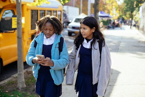 Free Cheerful multiracial girls classmates with backpacks browsing smartphone while walking together on sunny autumn street near yellow school bus Stock Photo