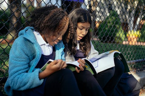 Focused multiracial girls pupils reading textbooks while sitting together on ground near net fence in autumn park