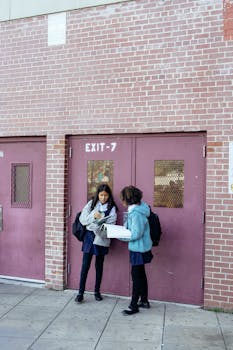 Full body girls classmates in warm jackets standing near masonry school building and reading textbooks attentively