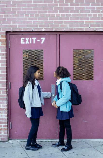 Side view full length multiethnic girls in casual outerwear standing near school door and having conversation