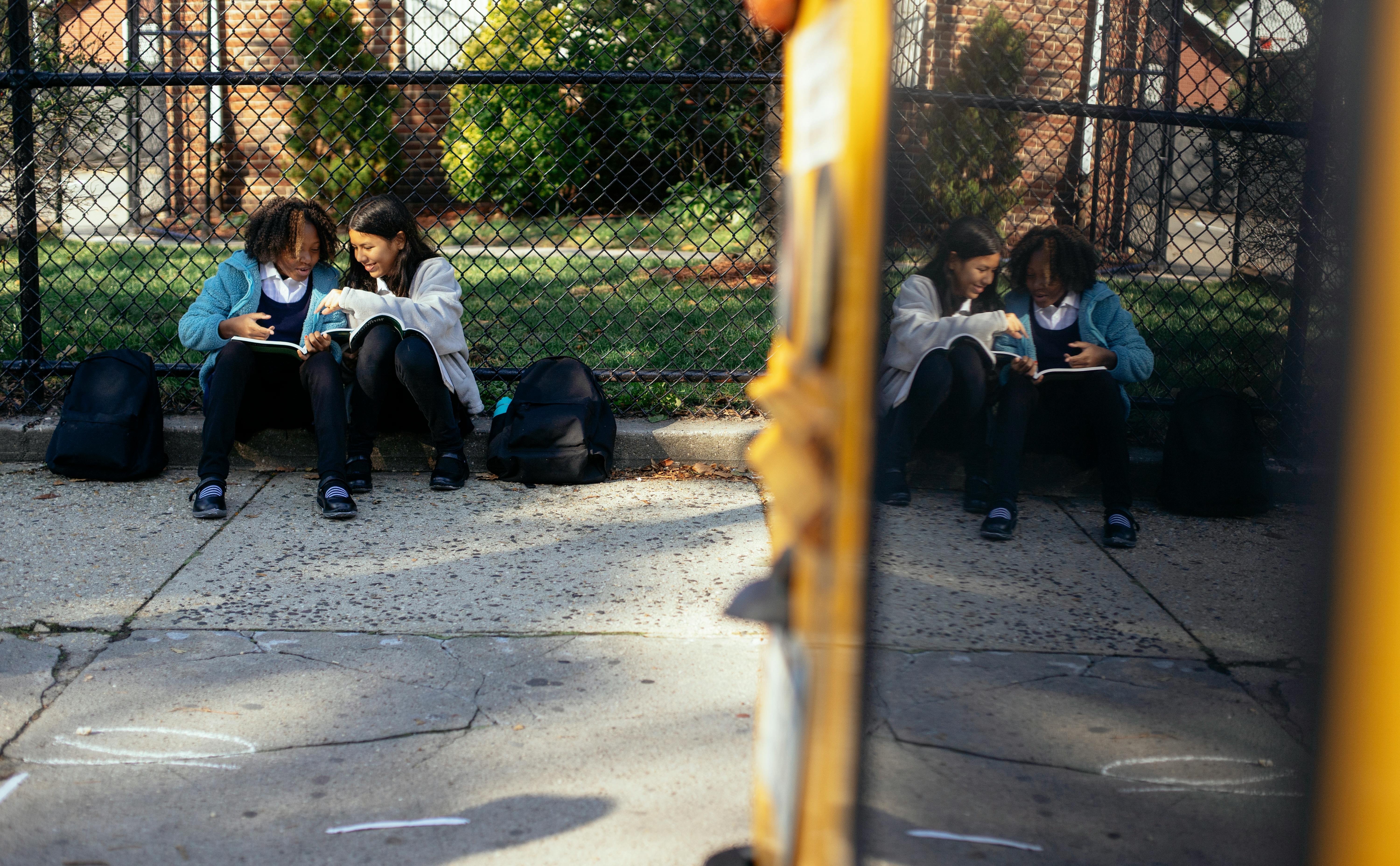 cheerful multiethnic schoolgirls sitting with textbooks near school fence