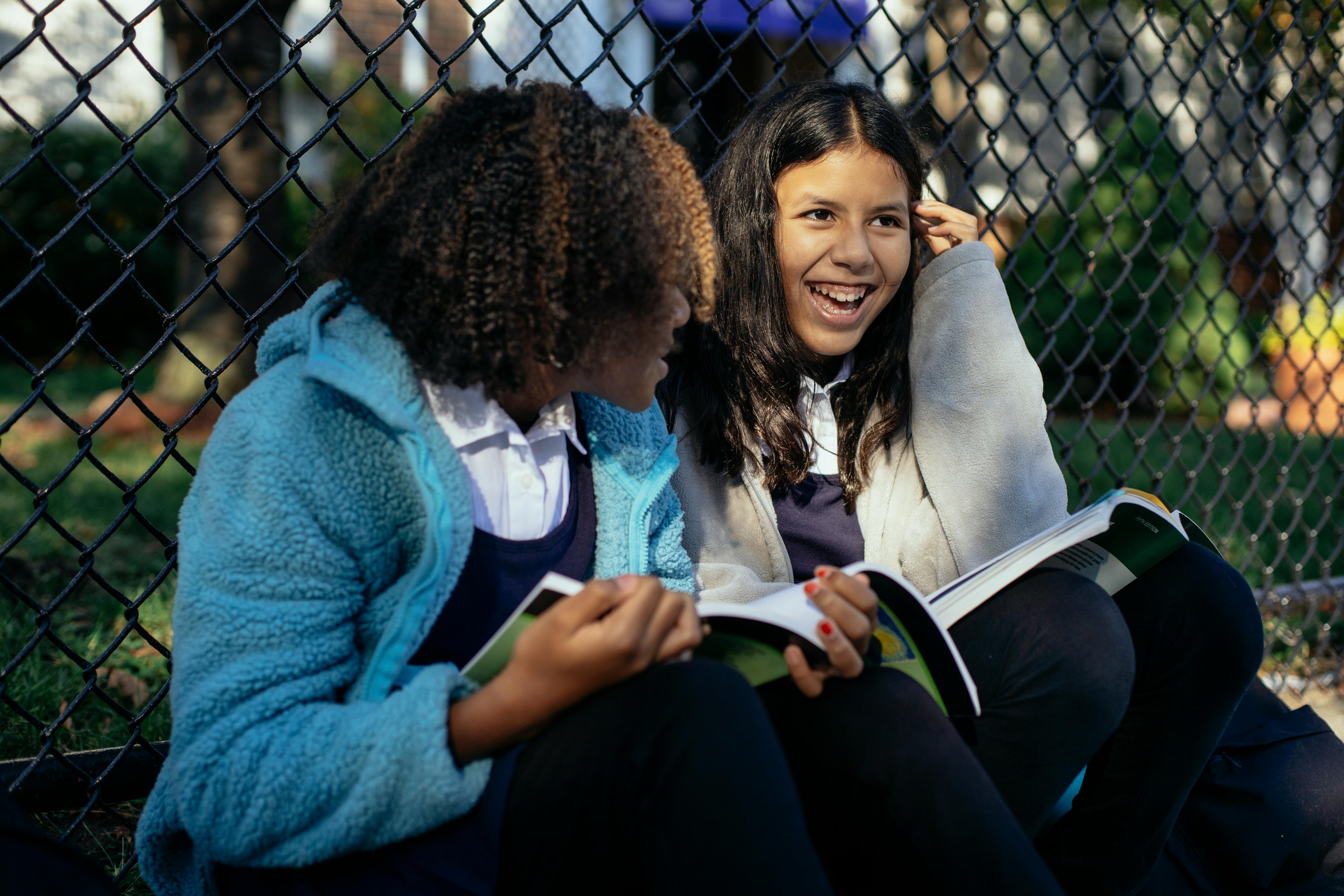 joyful multiethnic schoolgirls sitting with textbooks in autumn park