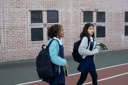 Two schoolgirls with backpacks walk outside a brick building, discussing school topics.