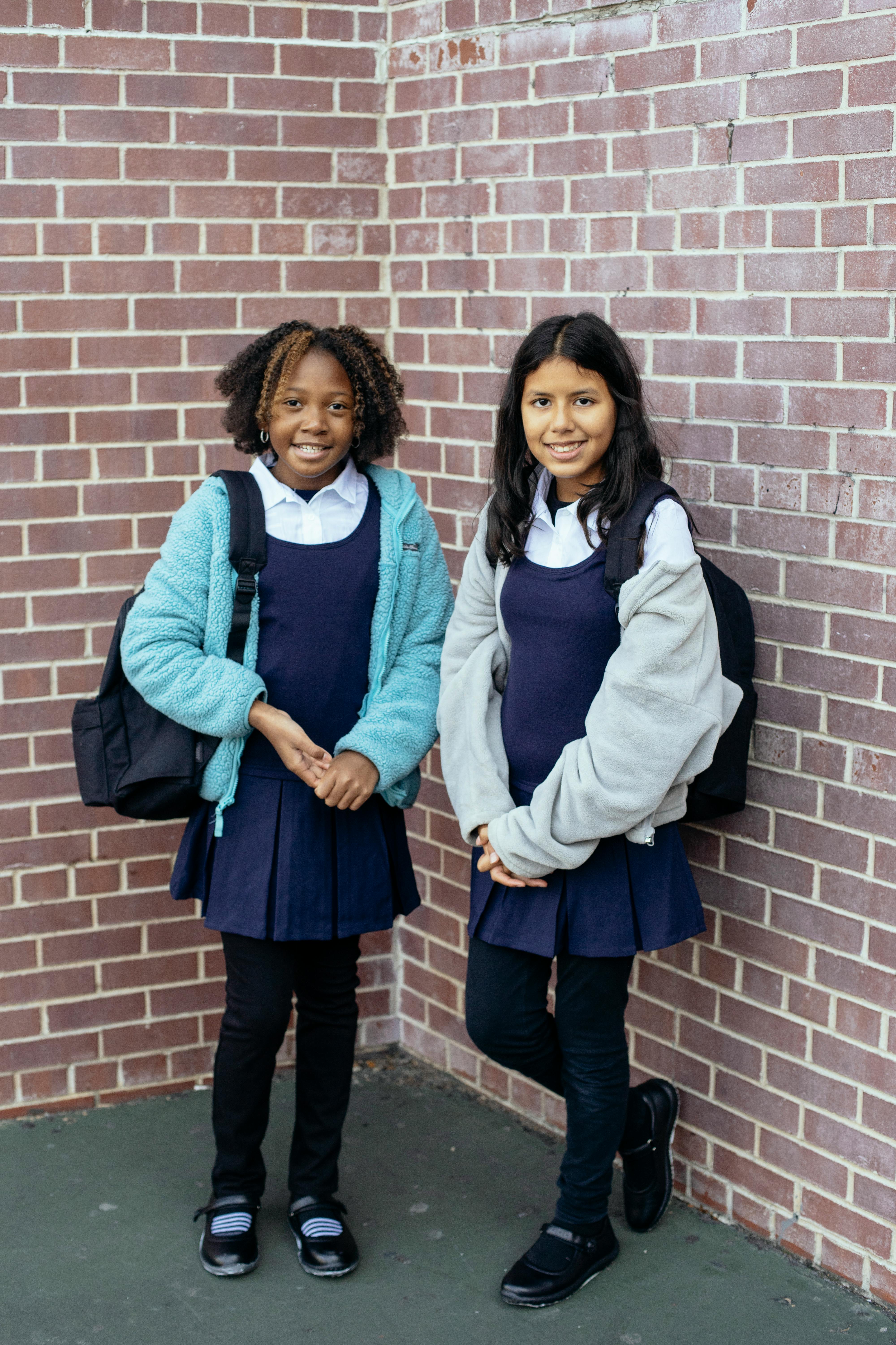 happy diverse schoolgirls in uniform smiling near brick wall