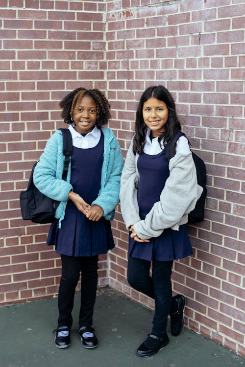 Free Happy diverse schoolgirls in uniform smiling near brick wall Stock Photo